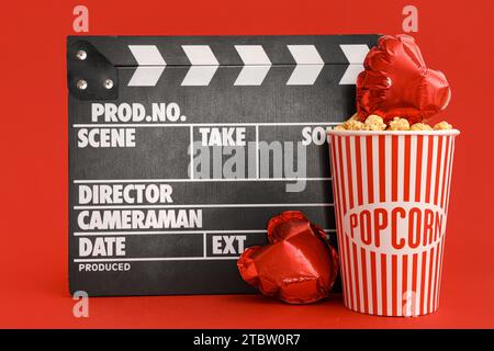 Movie clapper with bucket of popcorn and heart shaped air balloons on red background. Valentine's Day celebration Stock Photo