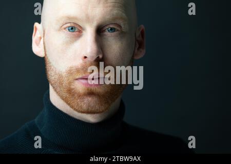 A man with a red beard looking straight and confident in black clothes in front of a black background in the camera Stock Photo