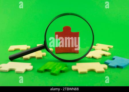 Colorful jigsaw puzzle pieces with magnifying glass focused on a single red corner piece in a conceptual image over green for search, investigation Stock Photo