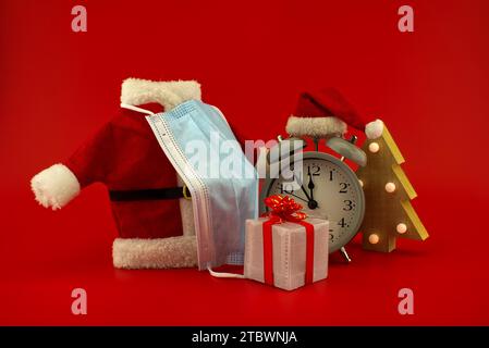 Christmas in pandemic concept still life with vintage alarm-clock, miniature santa costume and face mask, gift boxes on a festive red background Stock Photo
