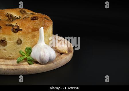 Round focaccia bread with olives, garlic and basil sprig over black background Stock Photo
