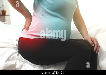 A pregnant woman is sitting on a bed touching her aching back (highlighted in red) Stock Photo