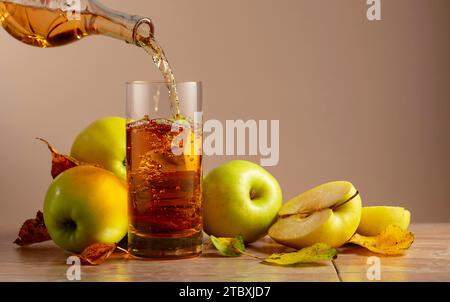 Apple juice is poured from a bottle into a glass. On a ceramic table ripe apples and dried-up leaves. Beige background with copy space. Stock Photo