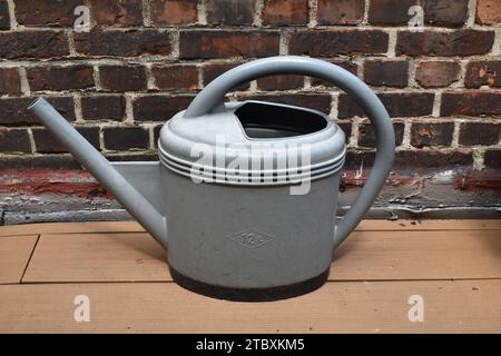 Grey plastic watering can with '12L' marking on light wooden floor in front of a dark red brick wall Stock Photo