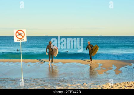 Two men walking into the ocean with their boards at Trigg Beach near a sign indicating the presence of rocks, Perth, Western Australia. Stock Photo