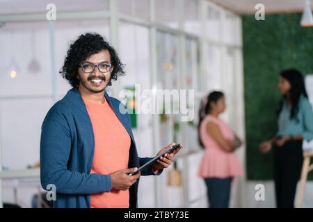 Confident young businessman at office standing by looking at camera - concept of IT professional, startup entrepreneur and corporate culture Stock Photo