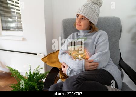 A woman in warm clothes sits in an armchair and hugs a lantern with candle. Low temperature in the house. Energy crisis concept.. Stock Photo