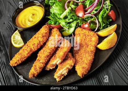 close-up of breaded fish fillet baked in oven served with yellow mustard, tortilla chips and fresh salad of lettuce, tomatoes, cucumber on black plate Stock Photo