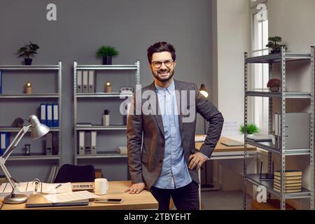 Portrait of smiling, happy, self-confident young businessman with glasses leaning on desk with stationery in spacious creative office. Stock Photo