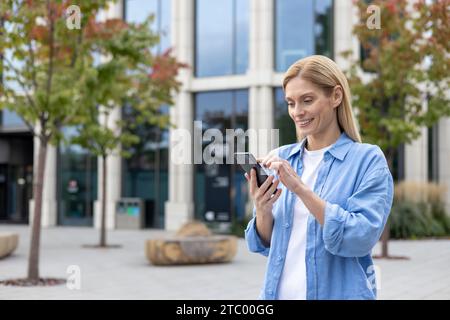 Mature woman with phone in hands walking in the city, a businesswoman in blue shirt holding smartphone in hands, reading online social networks, blonde smiling satisfaction is using an application. Stock Photo