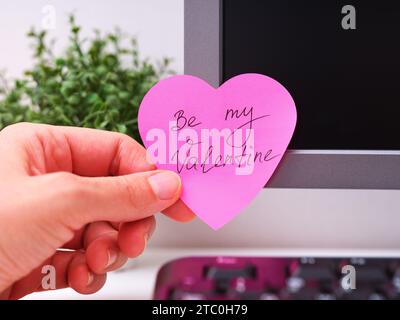 A person hand sticking a pink heart shaped paper note with words Be My Valentine on it on to a monitor at an office workplace Stock Photo