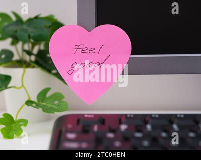 A pink heart shaped paper note with words Feel Good on it sticked on to a monitor at an office workplace. Stock Photo