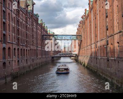 Tour boat heading down an avenue of red brick warehouses on Kehrweider fleet in the Speicherstadt district of Hamburg, Germany. Stock Photo