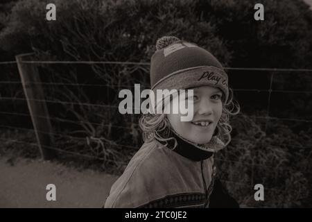 Young boy smiling at camera with long hair and wearing a beanie. Photo is black and white of boy walking along walking track on the Great Ocean Road. Stock Photo