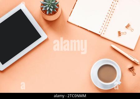 Overhead workplace with tablet computer, notepad, pen, coffee cup and succulent plant on peach background. Top view, flat lay, copy space. Stock Photo