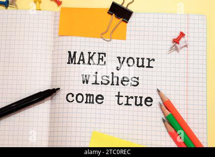 White notebook with inscription MAKE YOUR WISHES COME TRUE written in black pencil on a bright yellow background Stock Photo