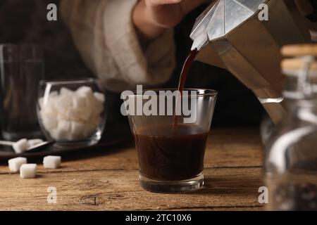 Woman pouring aromatic coffee from moka pot into glass at wooden table, closeup Stock Photo