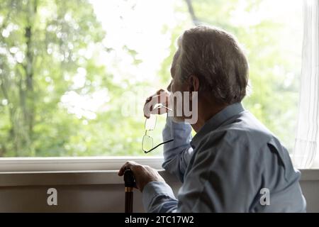 Pensive old man sit by window hold glasses walking stick Stock Photo