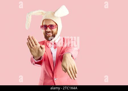Funny extravagant man in pink suit and hat with rabbit ears dancing on pink background. Stock Photo