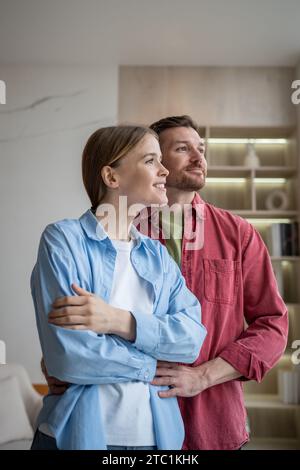 Gentle husband and tender wife standing embraced, looking at window, thinking of family, dreaming Stock Photo