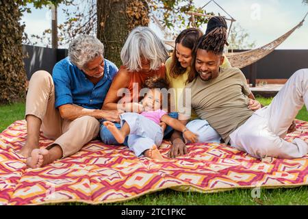 Multi-generational interracial family shares laughs and love on a colorful picnic blanket, embracing a cheerful outdoor moment. Stock Photo