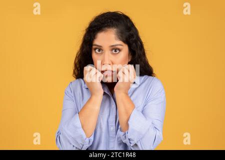 A worried young woman with curly hair wearing a blue shirt bites her nails and looks directly at the camera Stock Photo