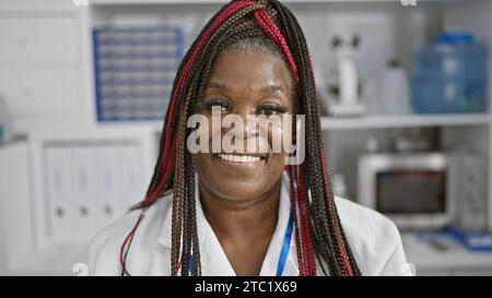 Confident african american woman scientist with braids, smiling beautifully in busy analytics chemistry lab, engrossed in medical research using micro Stock Photo