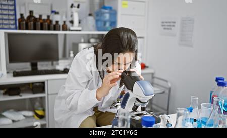 Dedicated young hispanic woman scientist, radiating beauty, immersed in deep concentration analyzing samples under her microscope at the laboratory Stock Photo