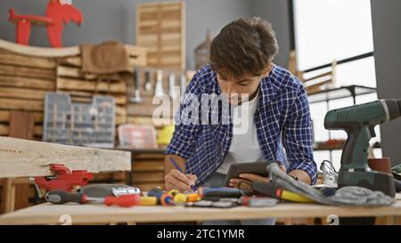Diligent young arabian man jotting down ideas on touchpad in carpenter's workshop, sculpting his mastery in woodwork Stock Photo