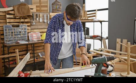 Handsome young arab man, a professional carpenter, concentrated on drawing a mark on a wood plank, fully engrossed in his carpentry work inside the wo Stock Photo