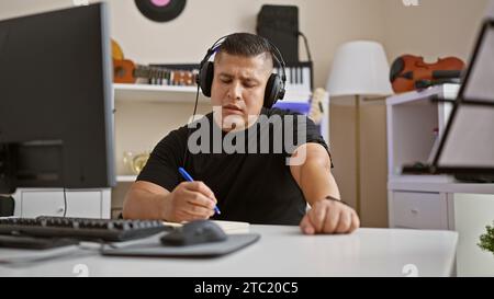 Talented young latin musician engrossed in crafting a soulful song at his cozy music studio, headphones on, effortlessly composing on his computer. Stock Photo