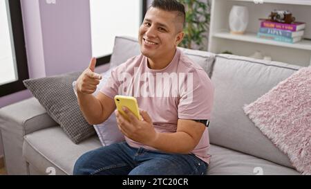 Cheerful young latin man enjoys typing a message, pointing at his smartphone while sitting on his living room sofa at home, happiness radiating from h Stock Photo