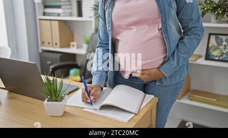 Expecting joy, young, hispanic working-mum-to-be massages belly while taking notes at office Stock Photo