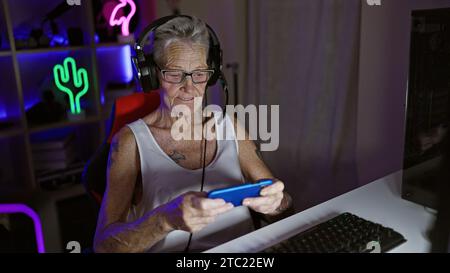 Cool grey-haired senior woman streamer rocks the gaming world, playing video game on smartphone, immersed in virtual entertainment, middle-aged but di Stock Photo
