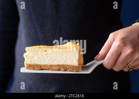 A woman holding a deep filled indulgent single portion of New York style Cheesecake on a cake slice Stock Photo