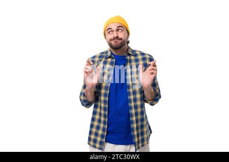 young doubtful european brunette man with a beard and mustache dressed in a yellow cap and a blue shirt shrugs his shoulders on a white background Stock Photo