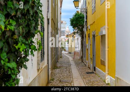 A narrow cobblestone residential alley in the historic old town of Cascais, Portugal, with the tower of Church of Our Lady of the Navigators in view. Stock Photo