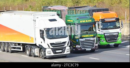 Close up front view group of three haulier businesses operating hgv supply chain transport lorries trucks overtaking four lane M25 motorway England UK Stock Photo