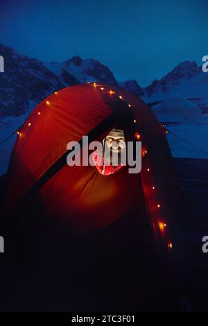 Man in red jacket sitting in camp tent with glowing Christmas glow garland lights in the winter mountains at night time Stock Photo