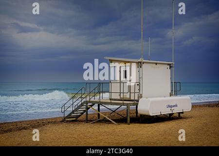 A gloomy, dramatic scene depicts a white lifeguard station amidst an incoming storm and waves breaking on a beach. Stock Photo