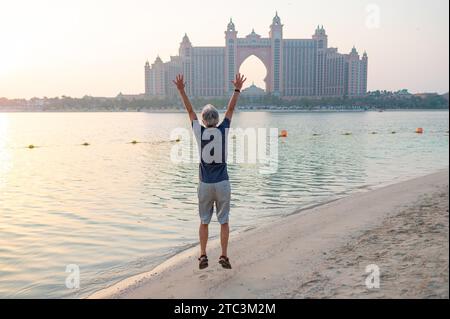 Senior caucasian man jumping high at Palm Jumeirah in Dubai, captured against the stunning backdrop of a sunset on the public beach Stock Photo