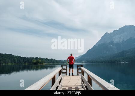 View from the shore of Lake Eibsee in Germany, with a hiker in a red shirt standing on a footbridge and looking into the distance. Stock Photo