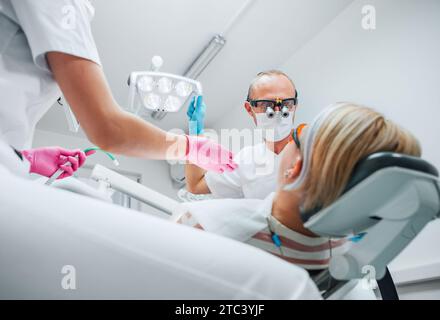 Dental clinic patient appointment. Dentist man in magnifying glasses pointing light. Young female assistant ready for teeth surgery with medical tools Stock Photo