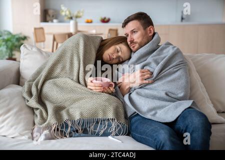 Sick sleepy woman feeling unwell, having high temperature, leaning on man shoulder with cup of tea Stock Photo