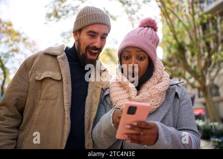 Young multiethnic couple using phone, looking at it surprised. People winning online gifts. Stock Photo