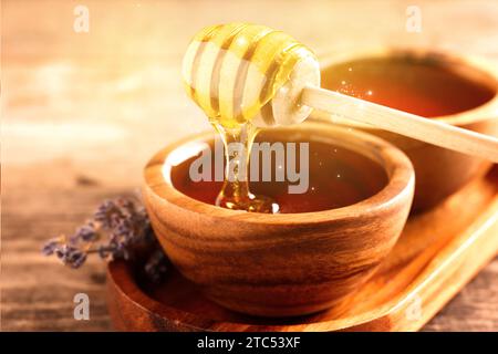 Natural honey dripping from dipper into wooden bowl on table under sunlight, closeup Stock Photo