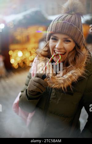 smiling modern woman in green coat and brown hat at the winter fair in the city with candy cane. Stock Photo