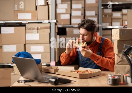 man in work clothes, a warehouse worker eats a meat pizza from fast food for lunch while sitting at the table. Stock Photo