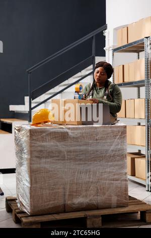 Stockroom worker preparing customer packages for delivery using adhesive tape, doing products quality control in warehouse. Stockroom supervisor working at online orders in storage room Stock Photo