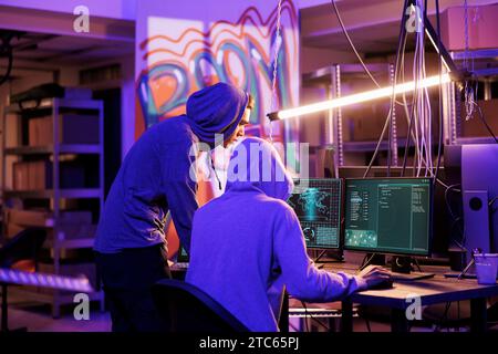 Internet criminals in hoods spying online using illegal spyware on computer. Hackers preparing ransomware attack, breaking law and planning phishing scam in abandoned warehouse Stock Photo
